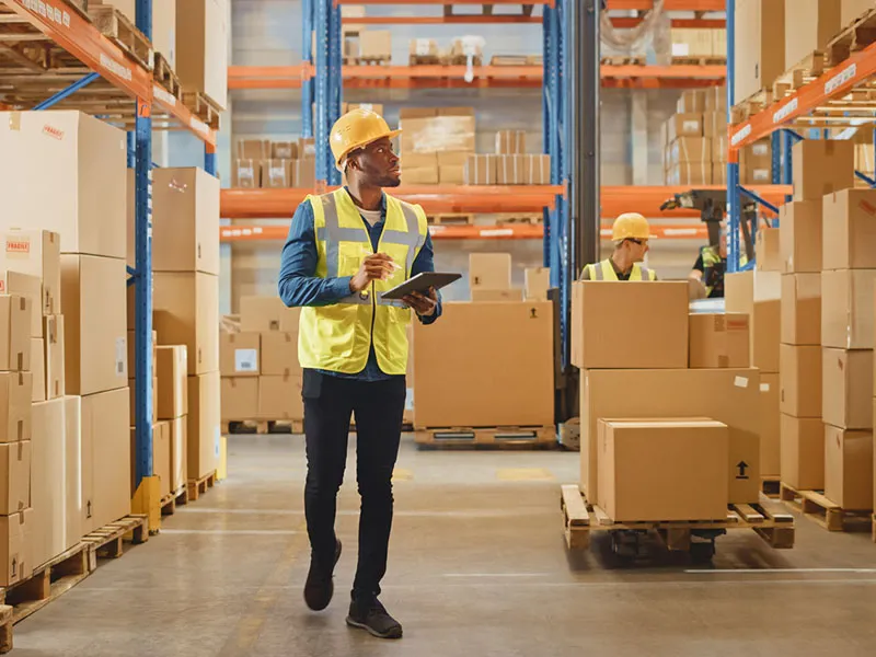 Handsome Male Worker Wearing Hard Hat Holding Digital Tablet Computer Walking Through Retail Warehouse full of Shelves with Goods. Working in Logistics and Distribution Center.